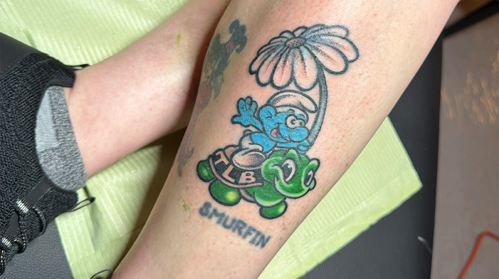Tattoo of a Smurf holding a large flower while riding a turtle with letters Smurfin under it